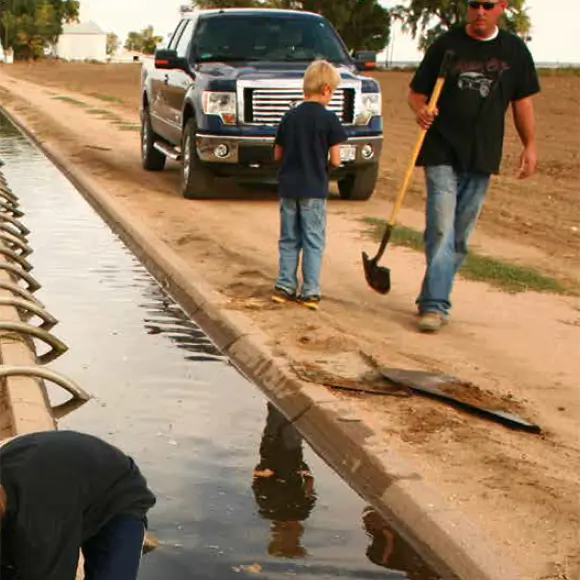 A man and a boy are near a rural irrigation ditch. A truck is parked on the dirt road. The man holds a shovel.