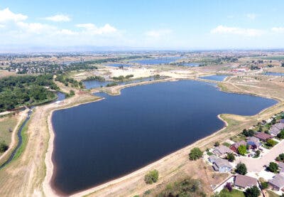Aerial view of a large pond or reservoir surrounded by homes, fields, and sparse vegetation under a clear blue sky.