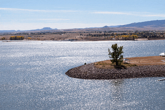 A lake with sunlight reflecting on the water, a small tree-covered island, and mountains in the background.