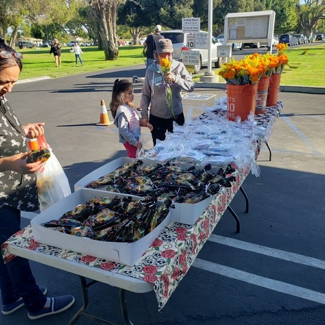 A table with packaged goods and marigold flowers is set up at an outdoor event, with people browsing nearby.