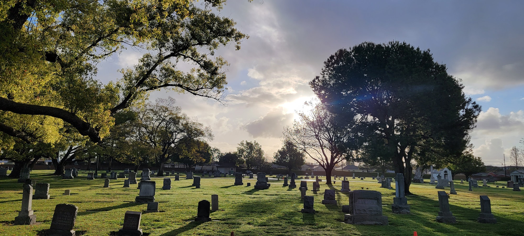 This picture contains a view of the grounds of the Anaheim Cemetery with a majestic sky of clouds and sun shining through the trees.