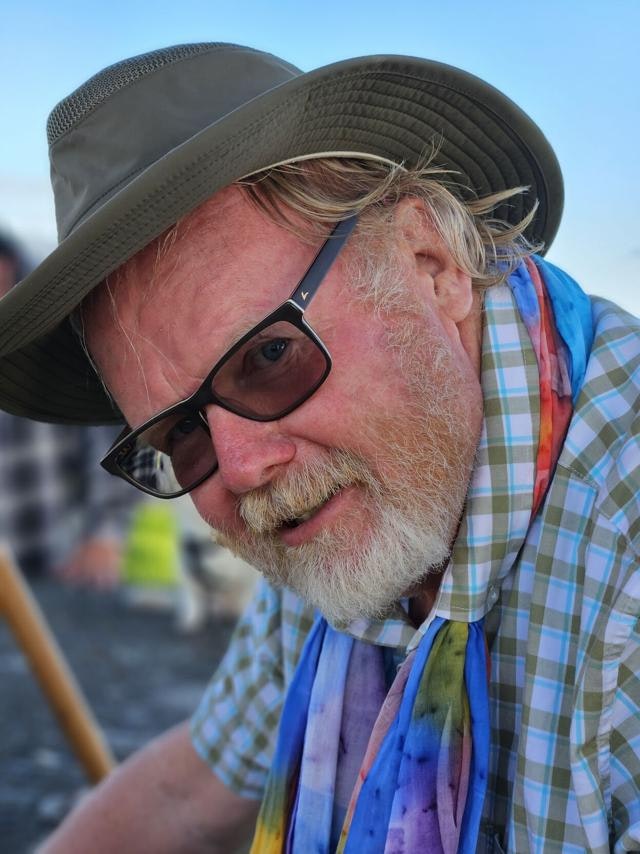 A man with a hat, glasses, and a colorful scarf, smiling in an outdoor setting.