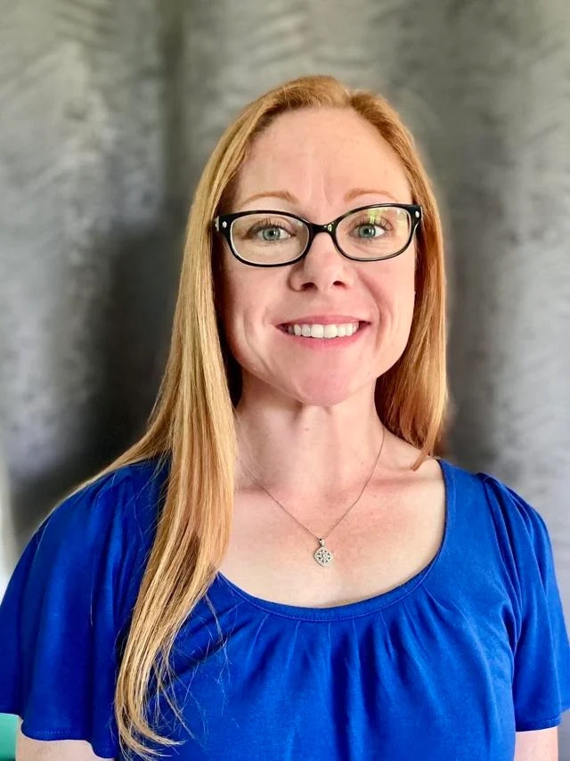 A person with glasses, long red hair, wearing a blue shirt and a necklace, smiling at the camera.