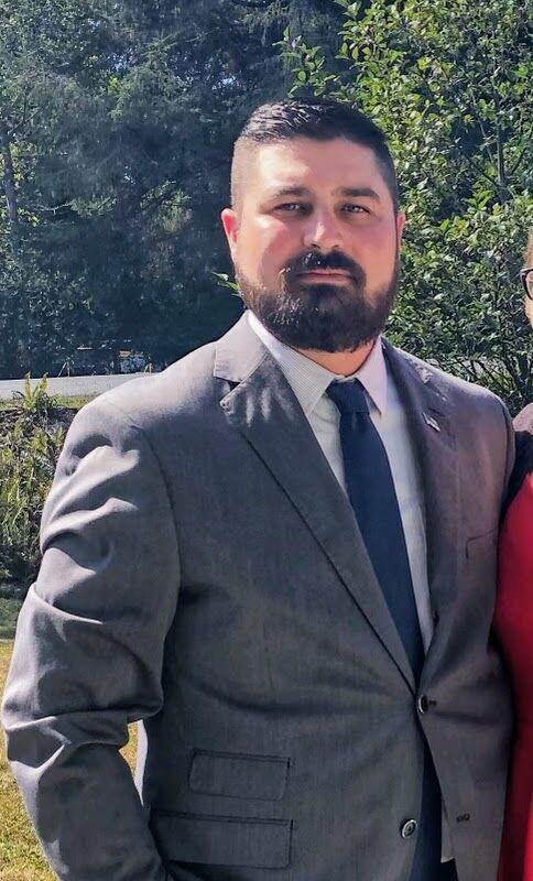 A man in a suit and tie stands outside, surrounded by greenery, looking at the camera.