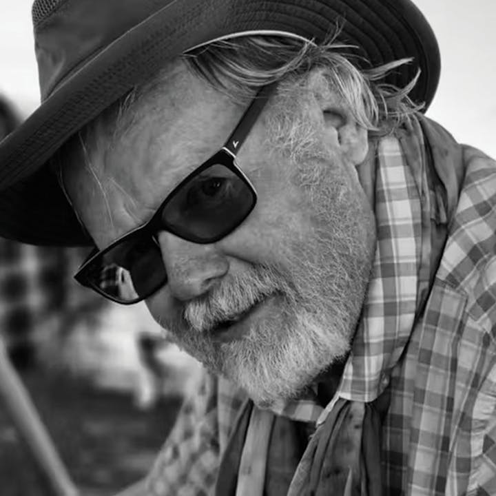 A person wearing sunglasses and a hat, with a beard and plaid shirt, looking at the camera in black and white.