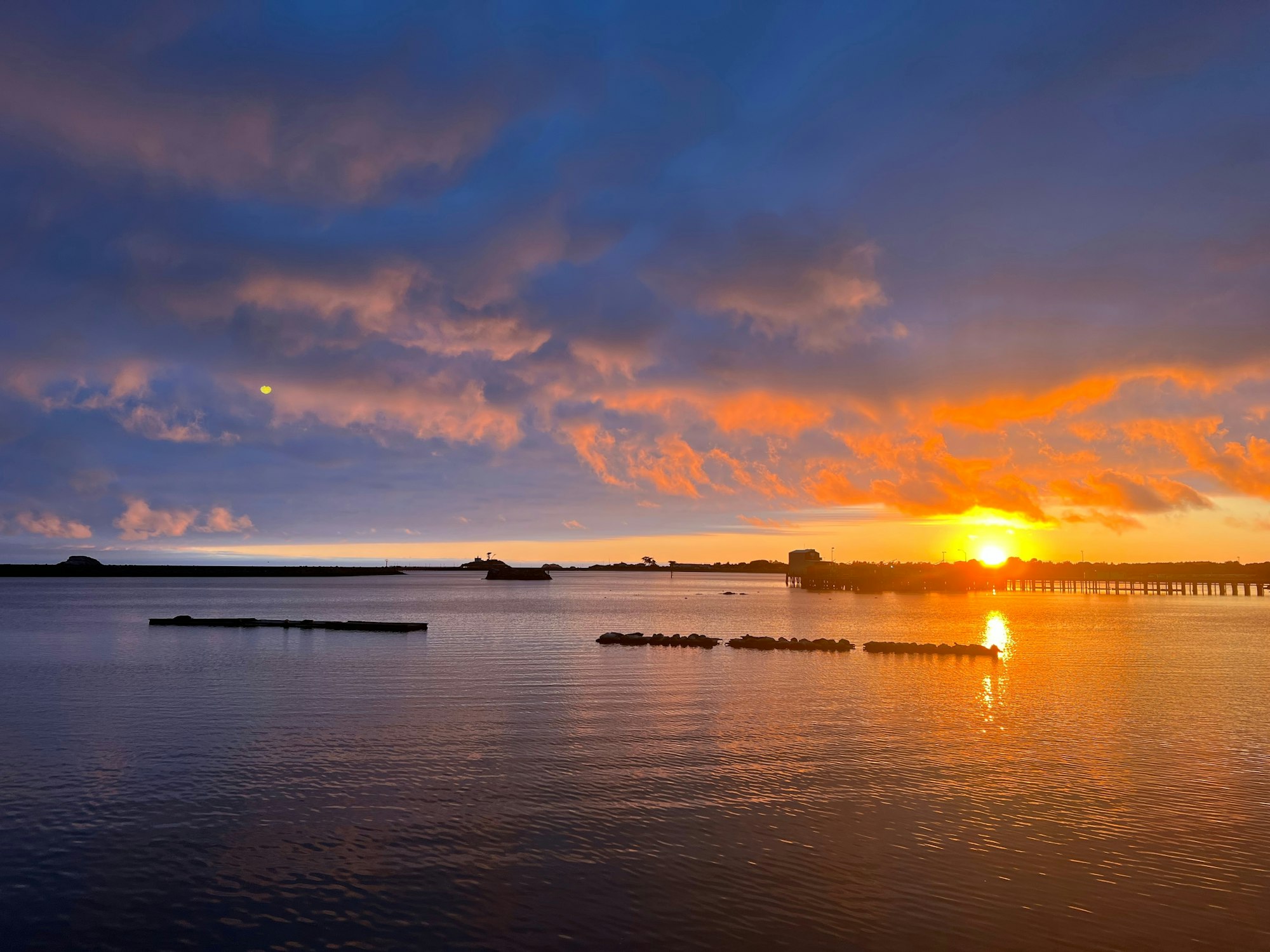 Sunset over a calm body of water with vibrant orange and purple clouds, silhouetted structures on the horizon.