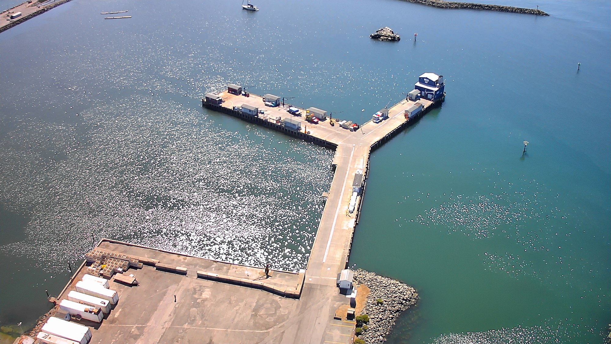 An aerial view of a dock area with a pier extension over calm water, surrounded by boats and structures along the shore.