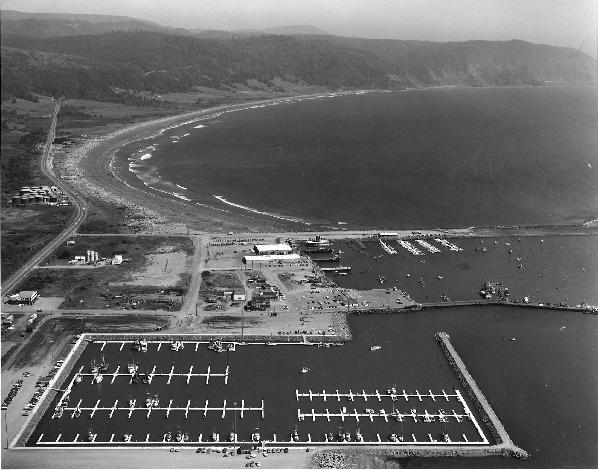 Aerial view of a coastal marina with boats, a curved beach, warehouses, roads, and surrounding forested hills.