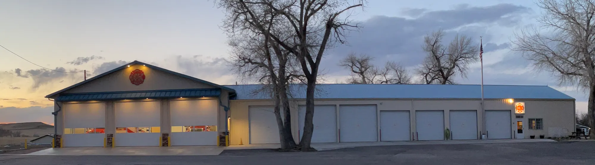 A large, well-lit building with multiple garage doors, set against a sunset sky and surrounded by sparse trees and grass.