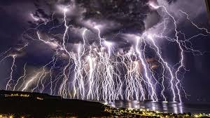 A dramatic sky filled with multiple lightning strikes over a body of water and cityscape.