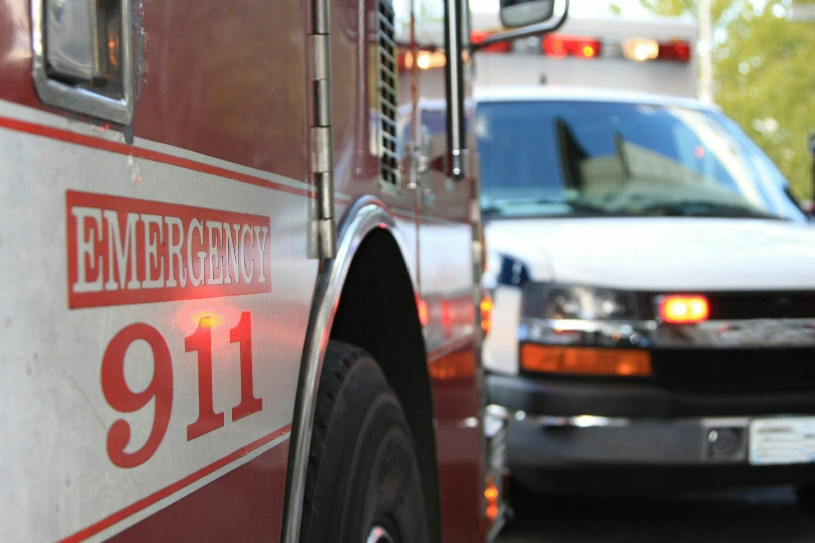 A close-up of a fire truck with "Emergency 911" on its side, blurred ambulance in the background.