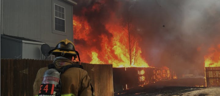 A firefighter observing a raging fire engulfing trees and a structure.