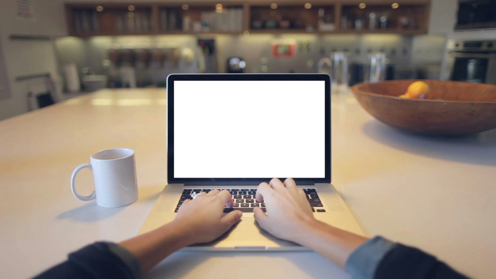 A person's hands typing on a laptop with a white mug on the table and a blurred kitchen background.