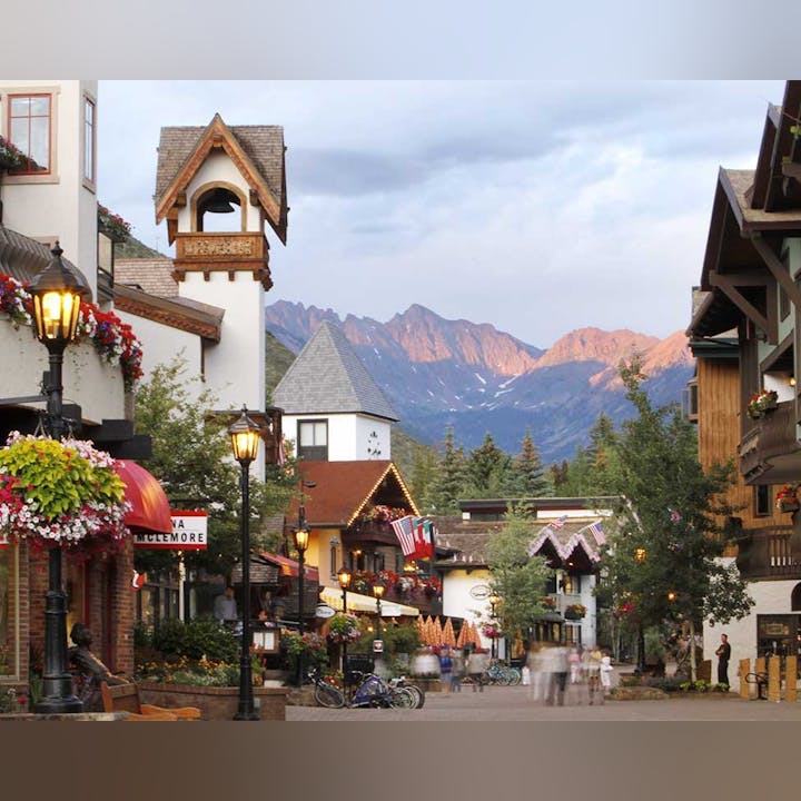 Charming town street lined with shops and flowering plants, with mountains in the background.