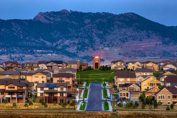 Suburban neighborhood with houses lined up, a street leading towards a clock tower, against a backdrop of mountains at dusk.