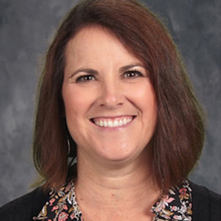 A smiling woman with shoulder-length hair, wearing a blouse with floral patterns and a dark jacket, against a mottled gray background.