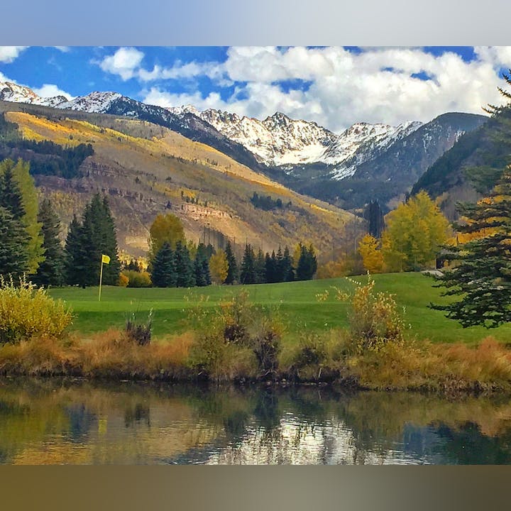 Snow-capped mountains, colorful autumn trees, a green golf course, and a pond reflecting the scenic landscape.