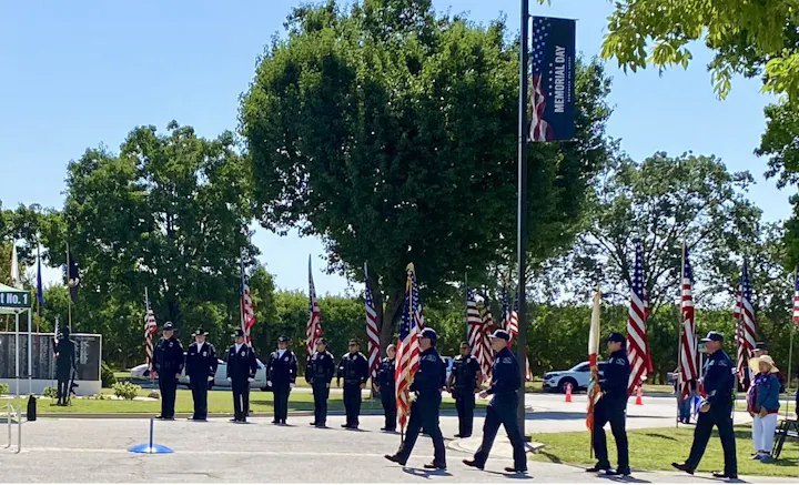 A group of uniformed personnel parades with flags during a Memorial Day event, surrounded by greenery and a commemorative site.
