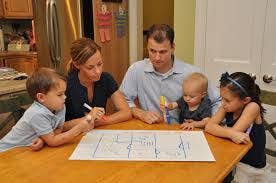 A family of five sits at a table, looking at a blueprint or plan, with two adults and three children in a kitchen setting.