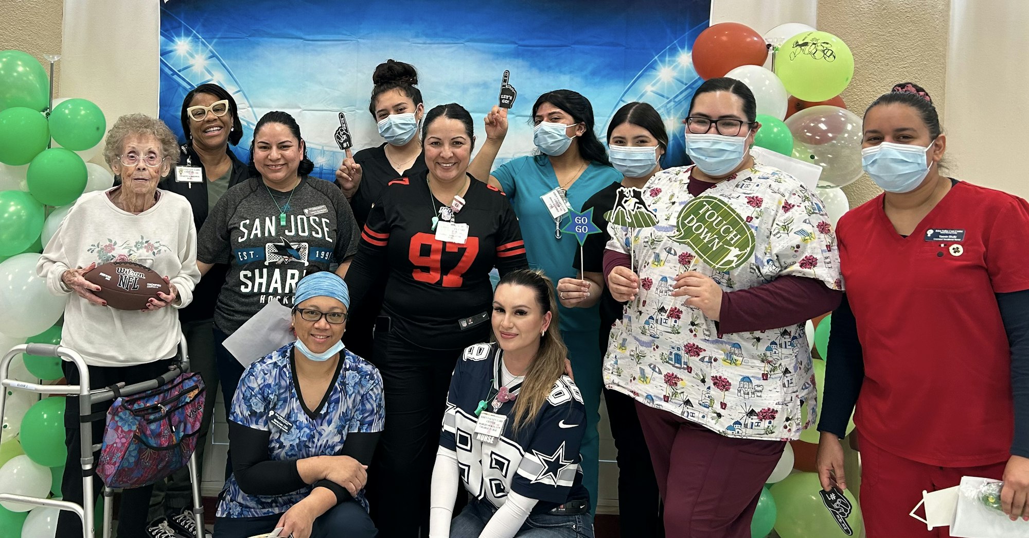 A group of smiling people, including healthcare workers in scrubs, celebrating with football-themed props and a backdrop.