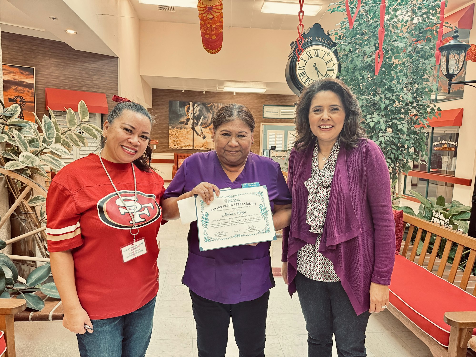 Three women smiling, middle one holding a certificate, indoor setting with a clock and plants.