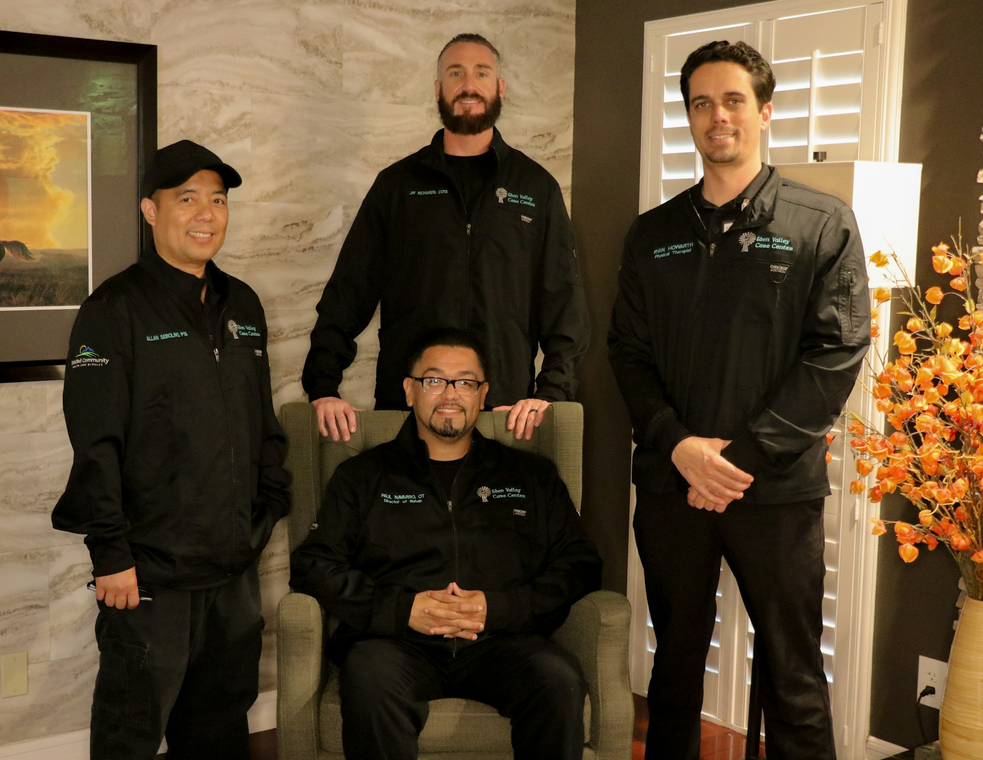 Four men in matching jackets posing in a room with a lamp and framed picture.