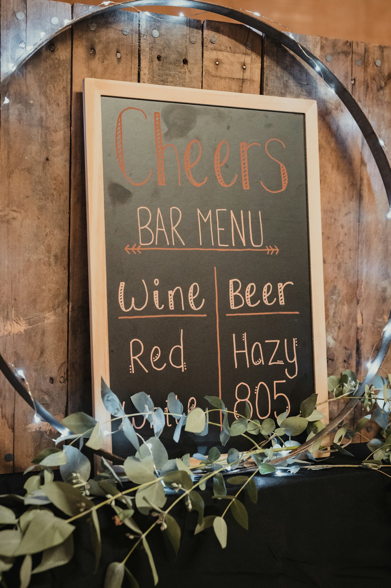 A wooden bar menu titled "Cheers" lists wine (red, white) and beer options (Hazy, 805), surrounded by greenery.