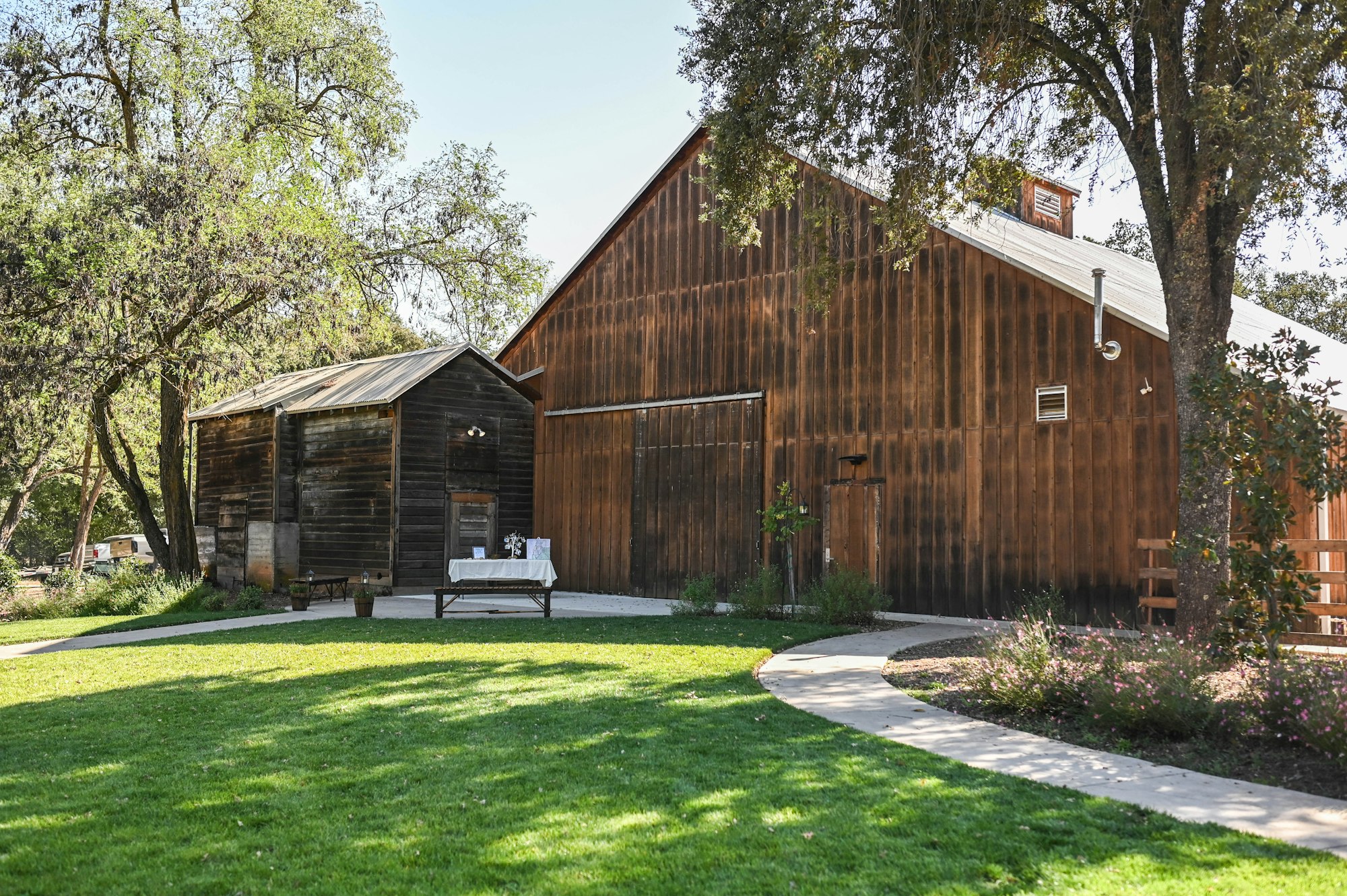 A scenic view of a rustic wooden barn and smaller shed, surrounded by green grass and trees, with a pathway.