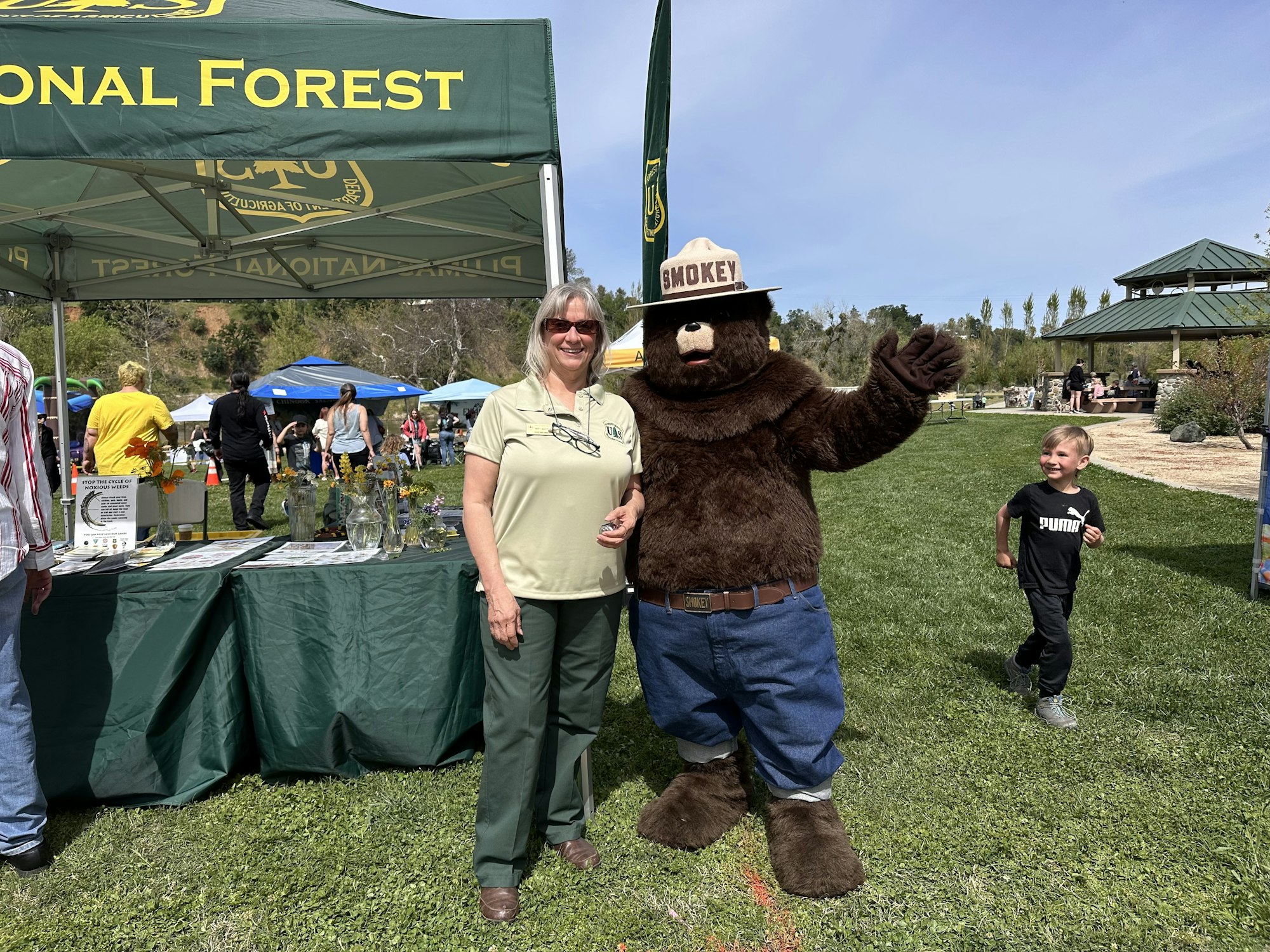 A woman and a Smokey Bear mascot stand under a "National Forest" tent at an outdoor event, with a child nearby.