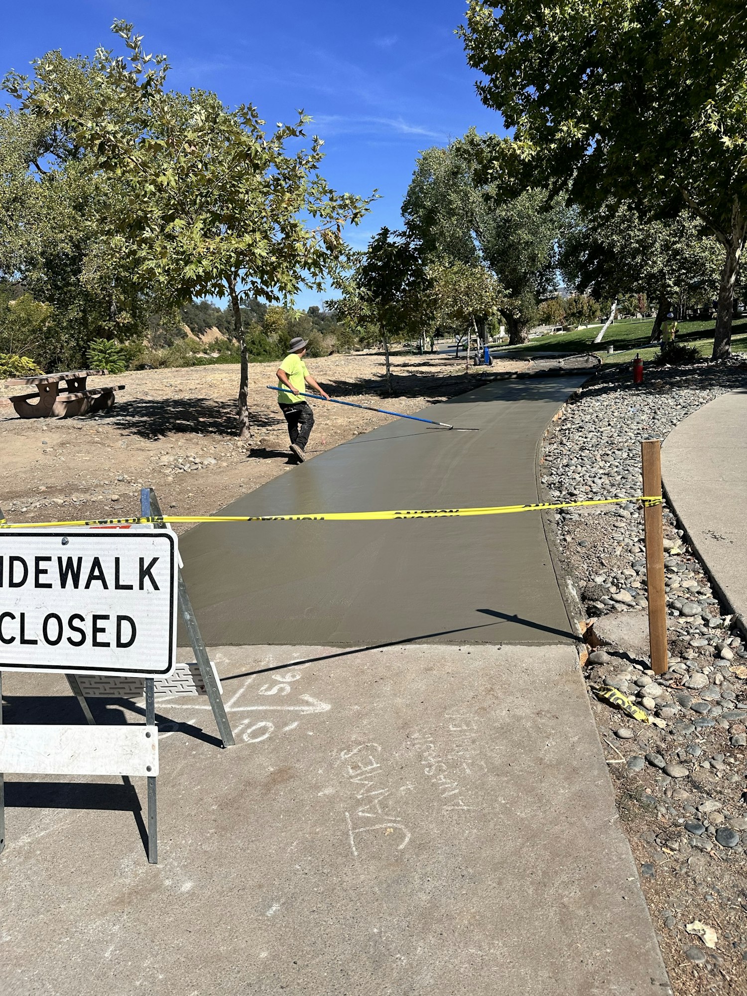 Freshly paved sidewalk with worker smoothing concrete, "Sidewalk Closed" sign, and caution tape.