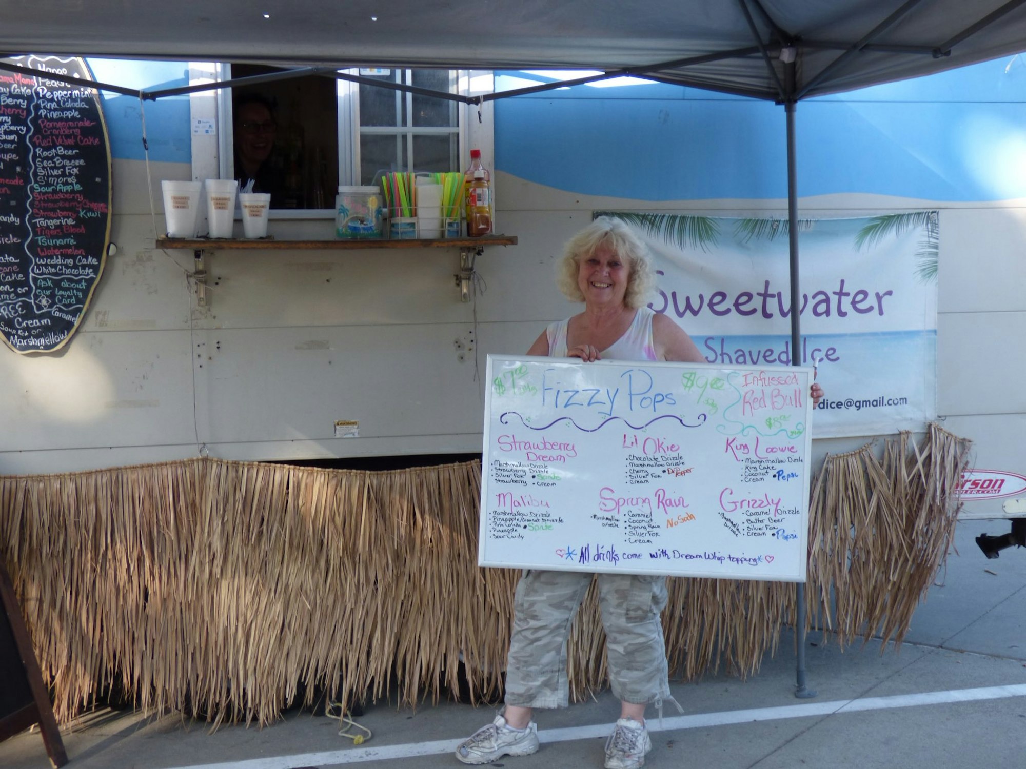 A woman holds a sign with menu items in front of the Sweetwater Shaved Ice stand, decorated with a tropical theme.