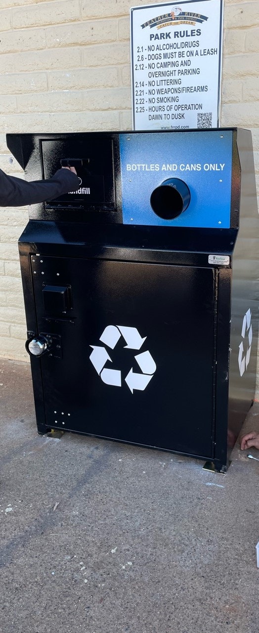 A recycling bin for bottles and cans, a list of park rules in the background, and a person's arm.