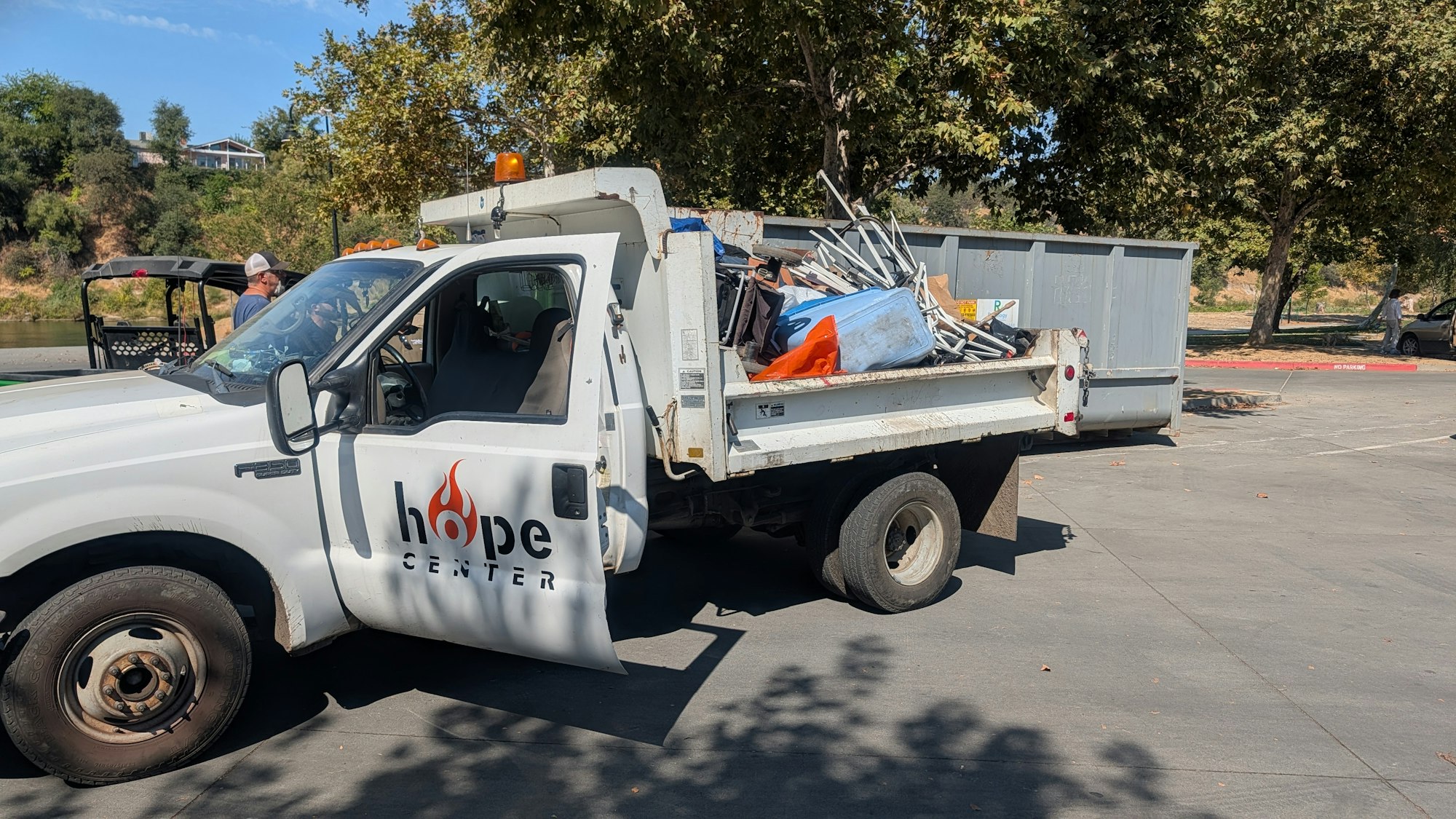 A white truck with "Hope Center" logo, filled with various items, parked in a sunny outdoor area with trees and people nearby.