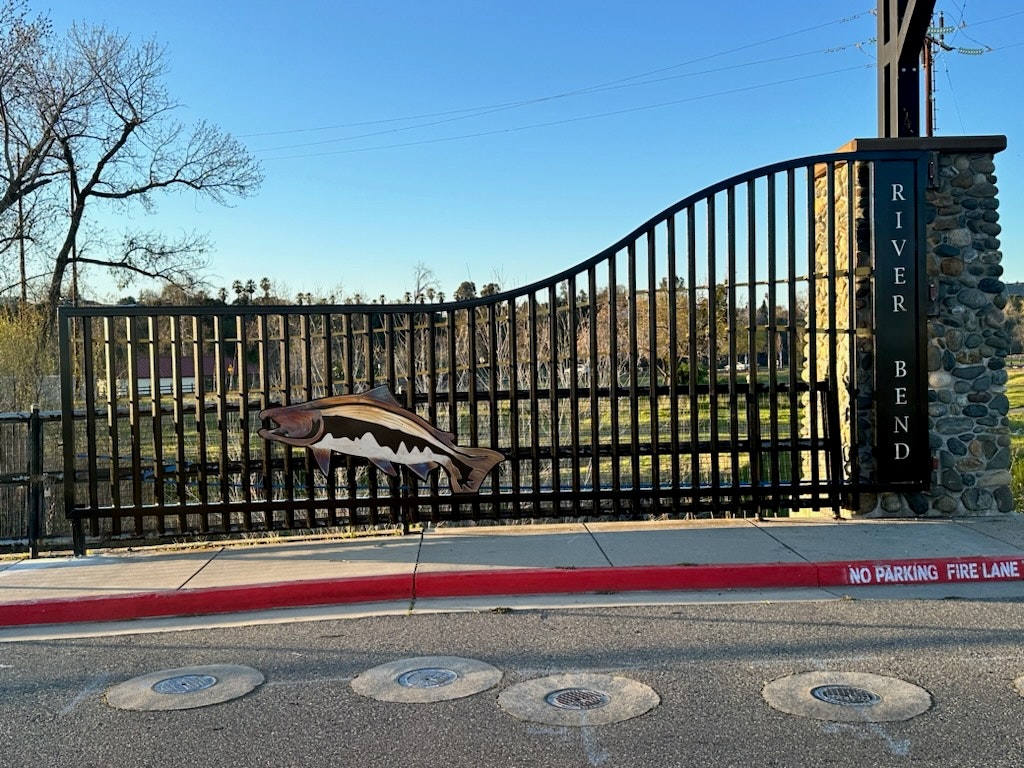 An ornate gate with a fish design labeled "RIVER BEND," next to a "NO PARKING FIRE LANE" curb.