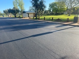 An empty parking lot, shadows on the ground, trees, and a small building in the background on a sunny day.