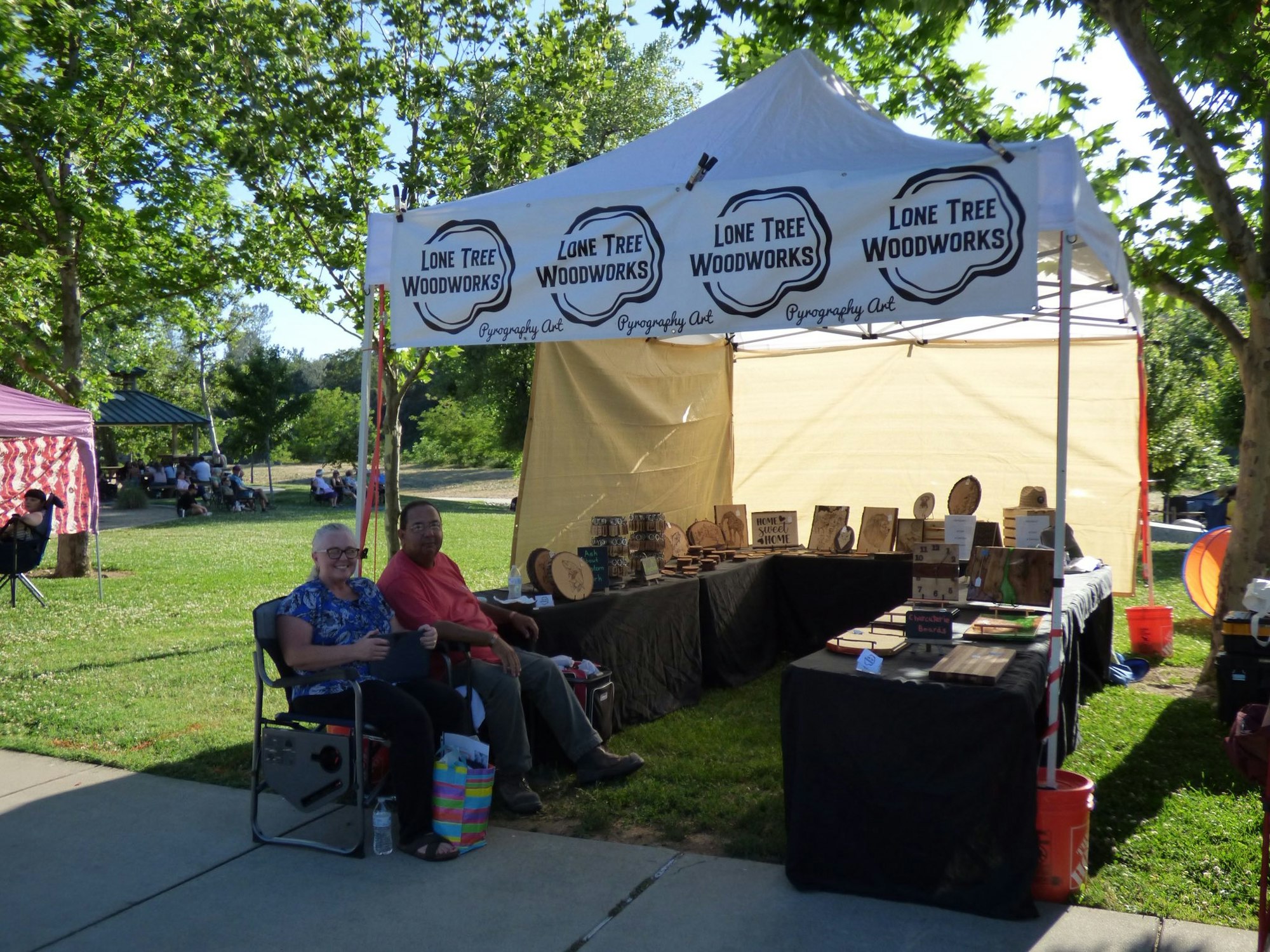 Outdoor market stall with woodworks display and two people sitting under a "Lone Tree Woodworks" tent.