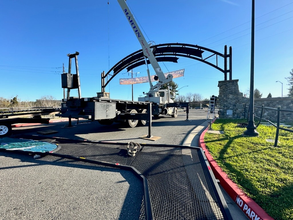 A crane assembling or disassembling a metal archway near a road with "NO PARKING" signs, clear skies, and greenery.