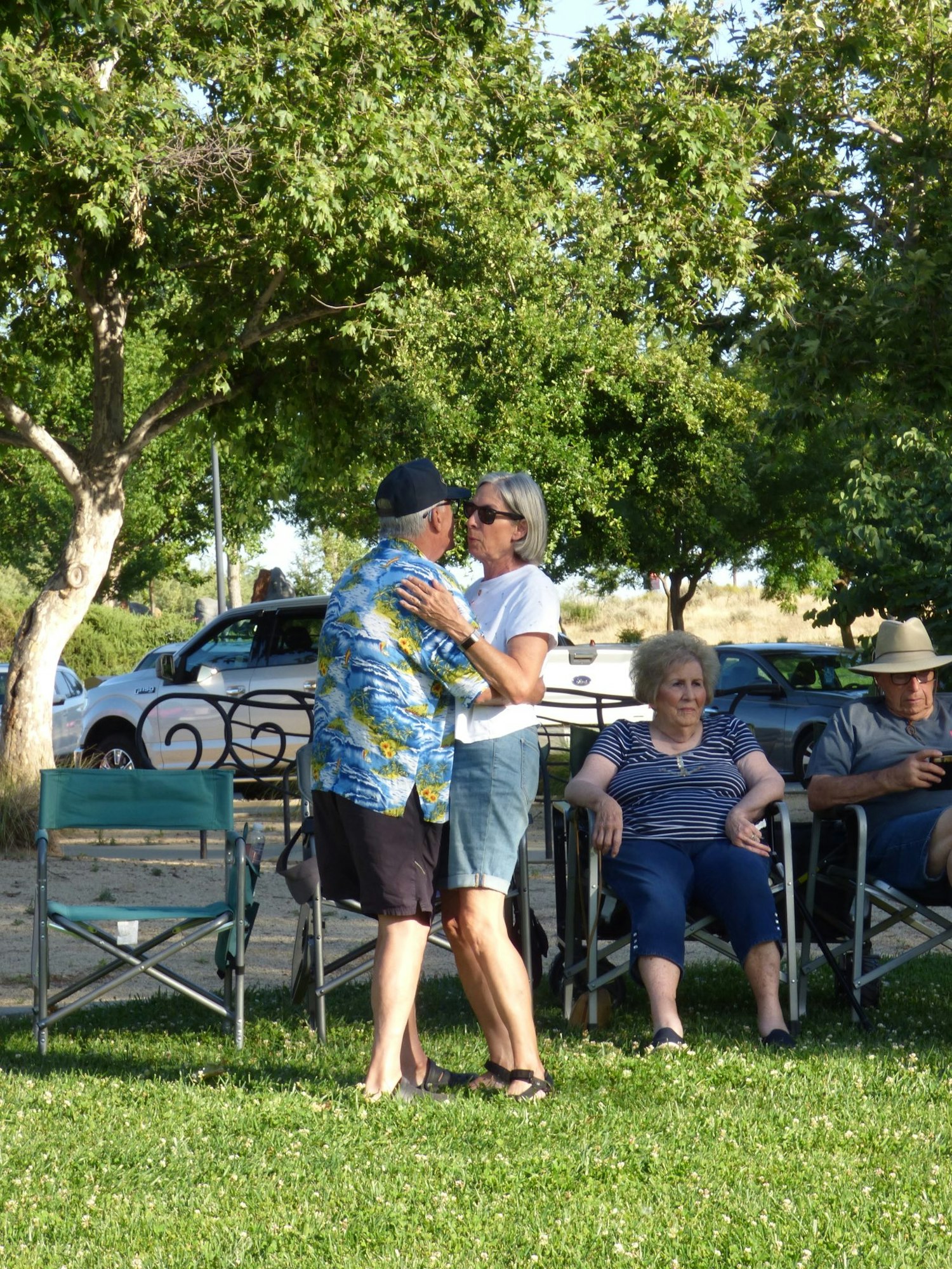 A couple dancing outdoors, two people seated nearby, with trees and a parked car in the background.