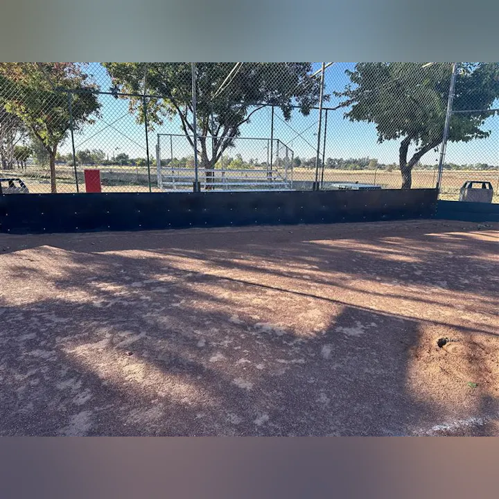 A view of a dusty baseball field with a black fence and trees in the background, likely at a park or sports complex.