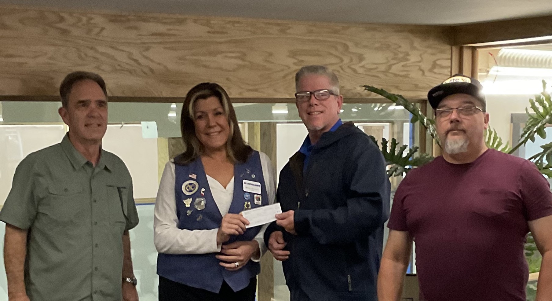 Four people posing, one giving a check to another, in an indoor setting with plants and wooden walls.