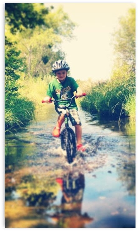 A child rides a bike through a shallow stream, splashing water while surrounded by greenery and trees.