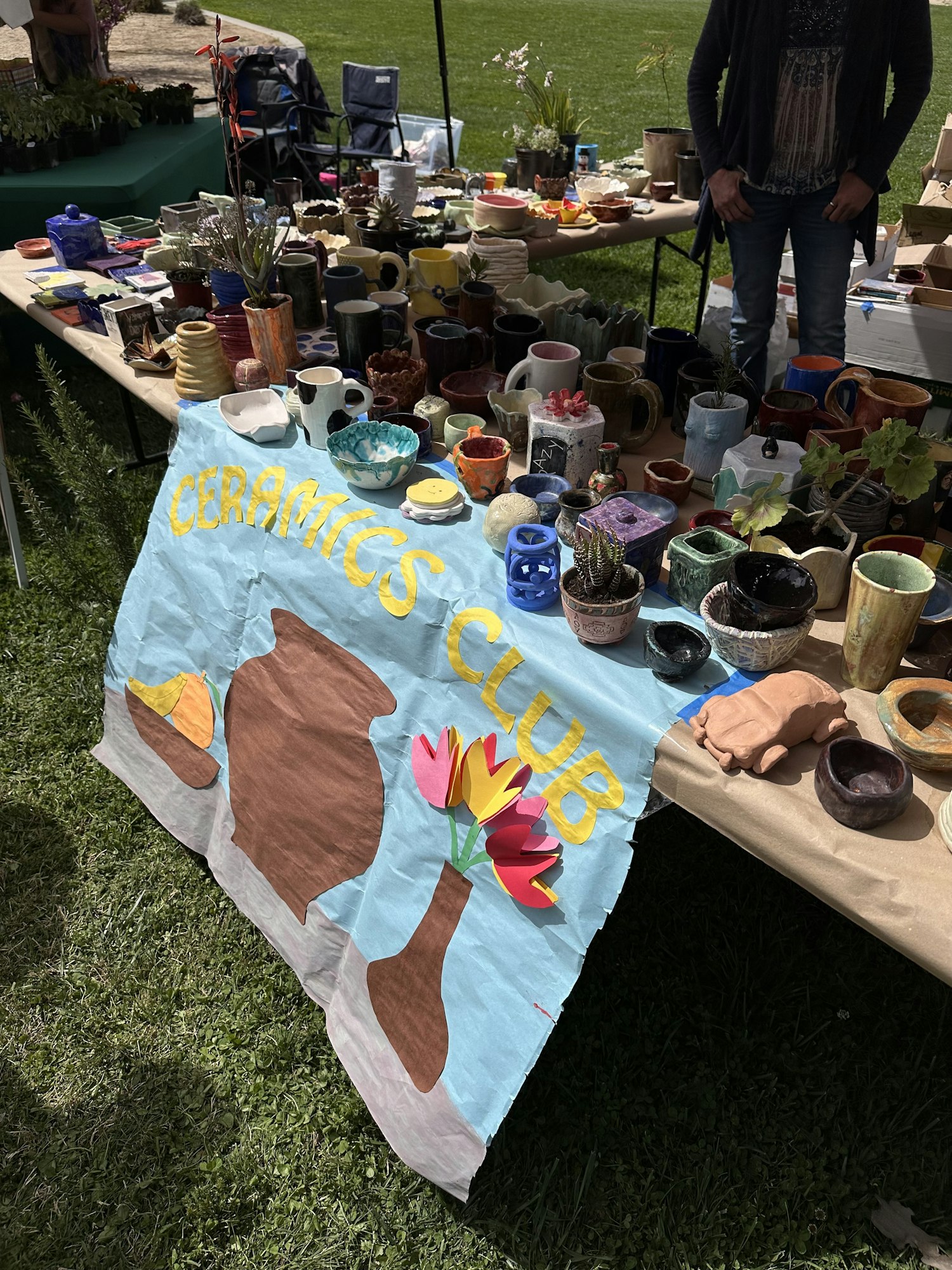 Ceramics Club table display with various handmade pottery and decorations set up on grass.