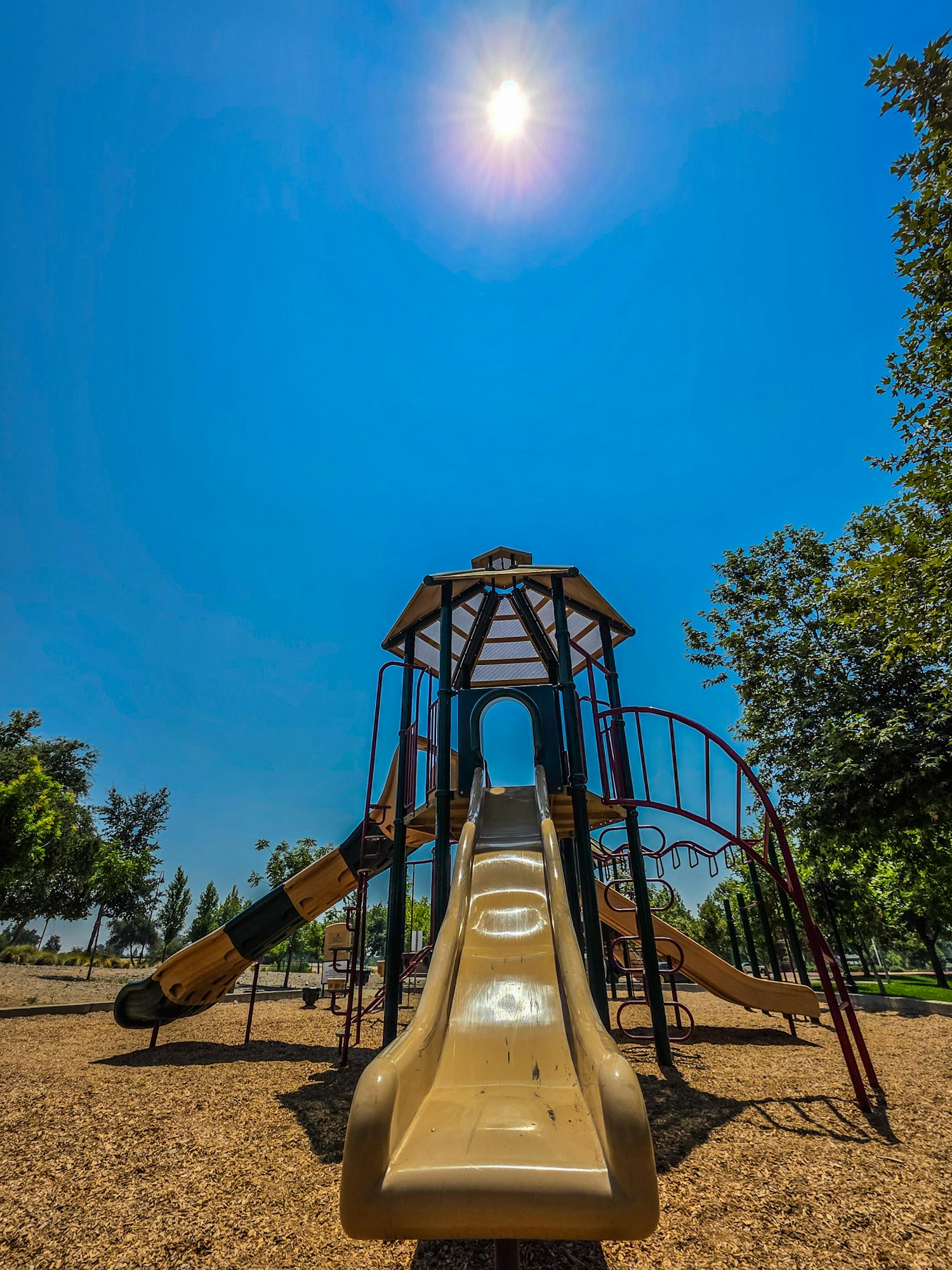 A playground slide under a bright sun with a clear blue sky and surrounding trees.