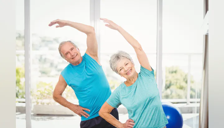 A senior couple enjoying a workout, stretching together in a bright room with a fitness ball in the background.
