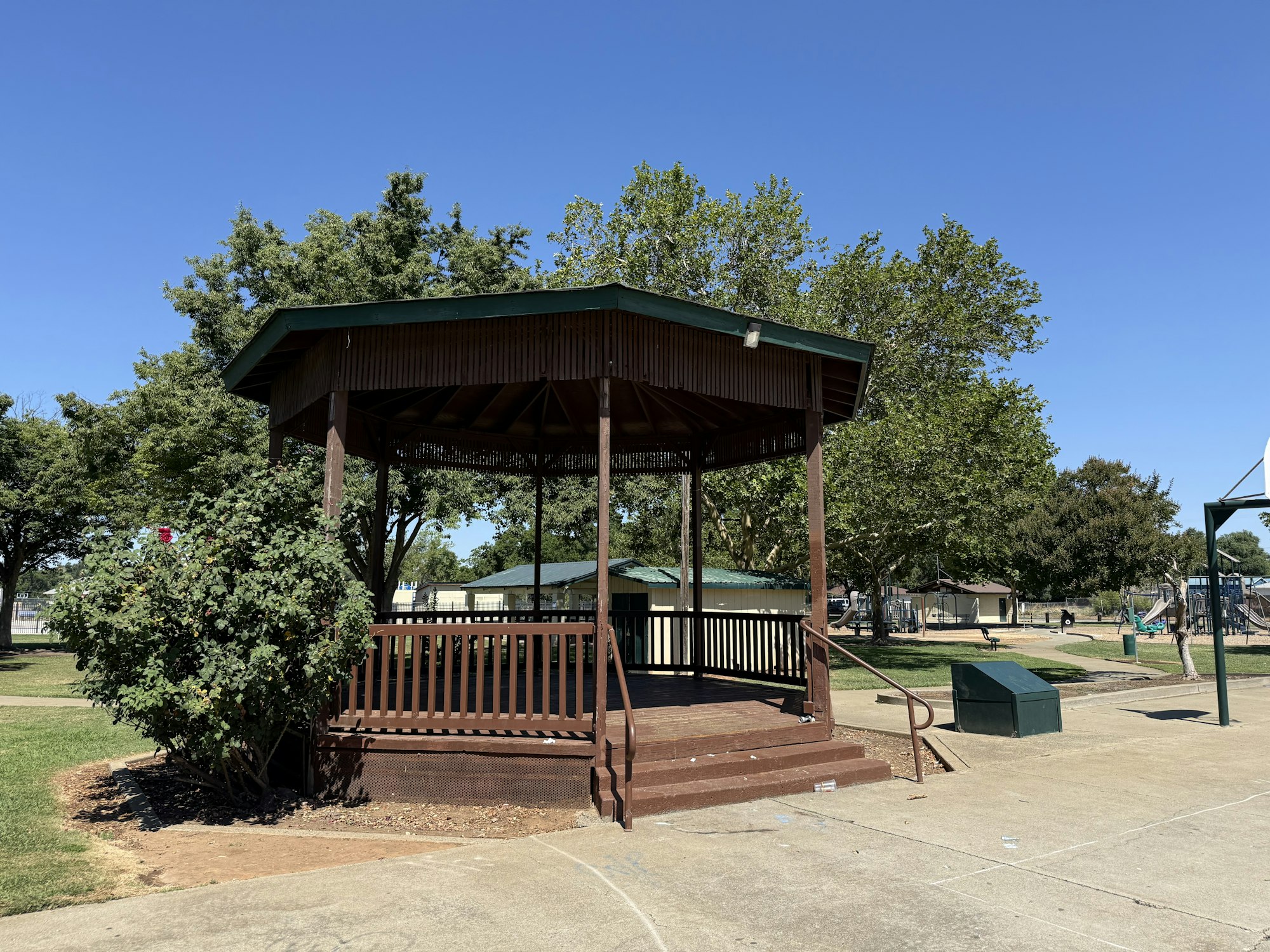 A wooden gazebo in a park, surrounded by greenery and a clear blue sky, with playground equipment in the background.