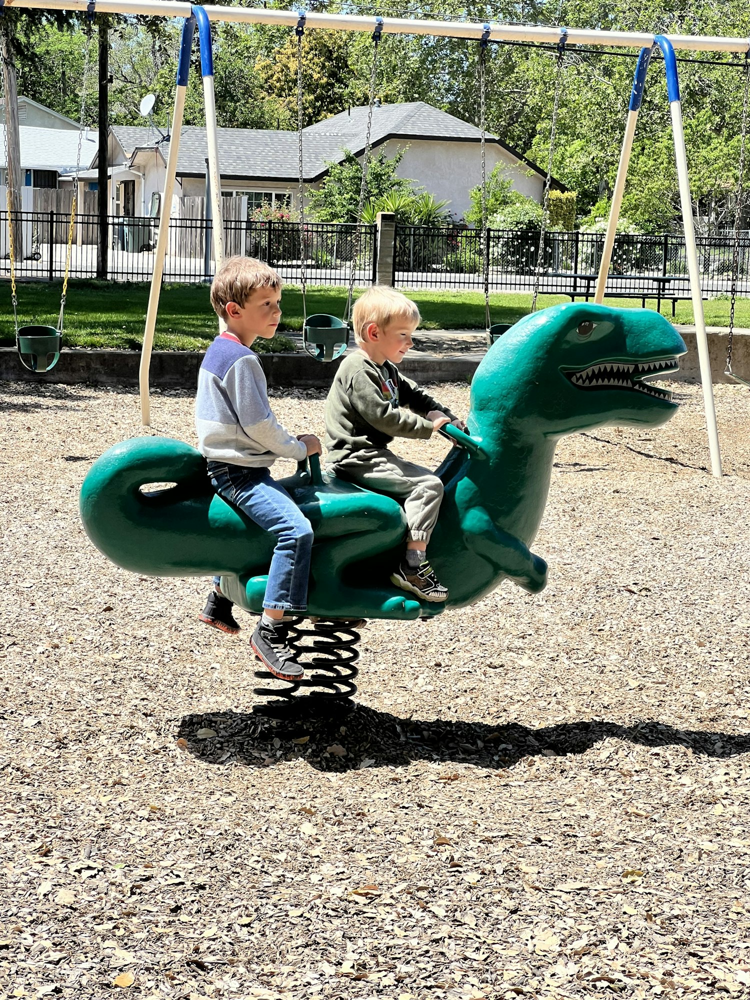 Two children are sitting on a green dinosaur spring rider in a playground.