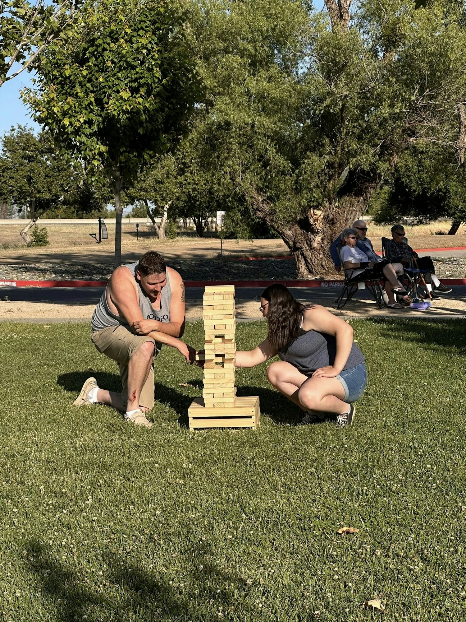 Two people are playing a large outdoor Jenga game on the grass, with onlookers seated in the background.