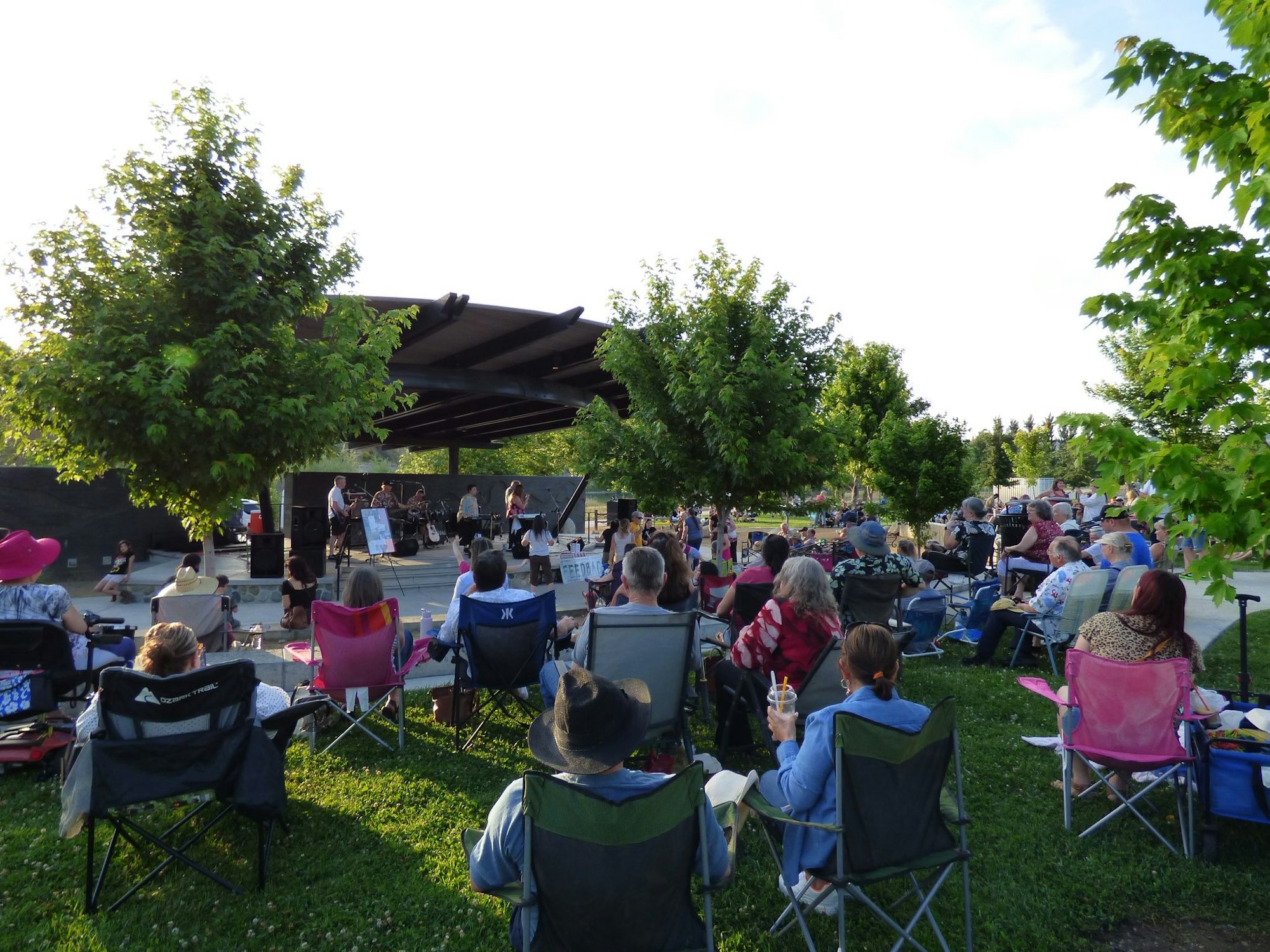 Outdoor concert with a band playing on stage and people sitting on lawn chairs, surrounded by trees.