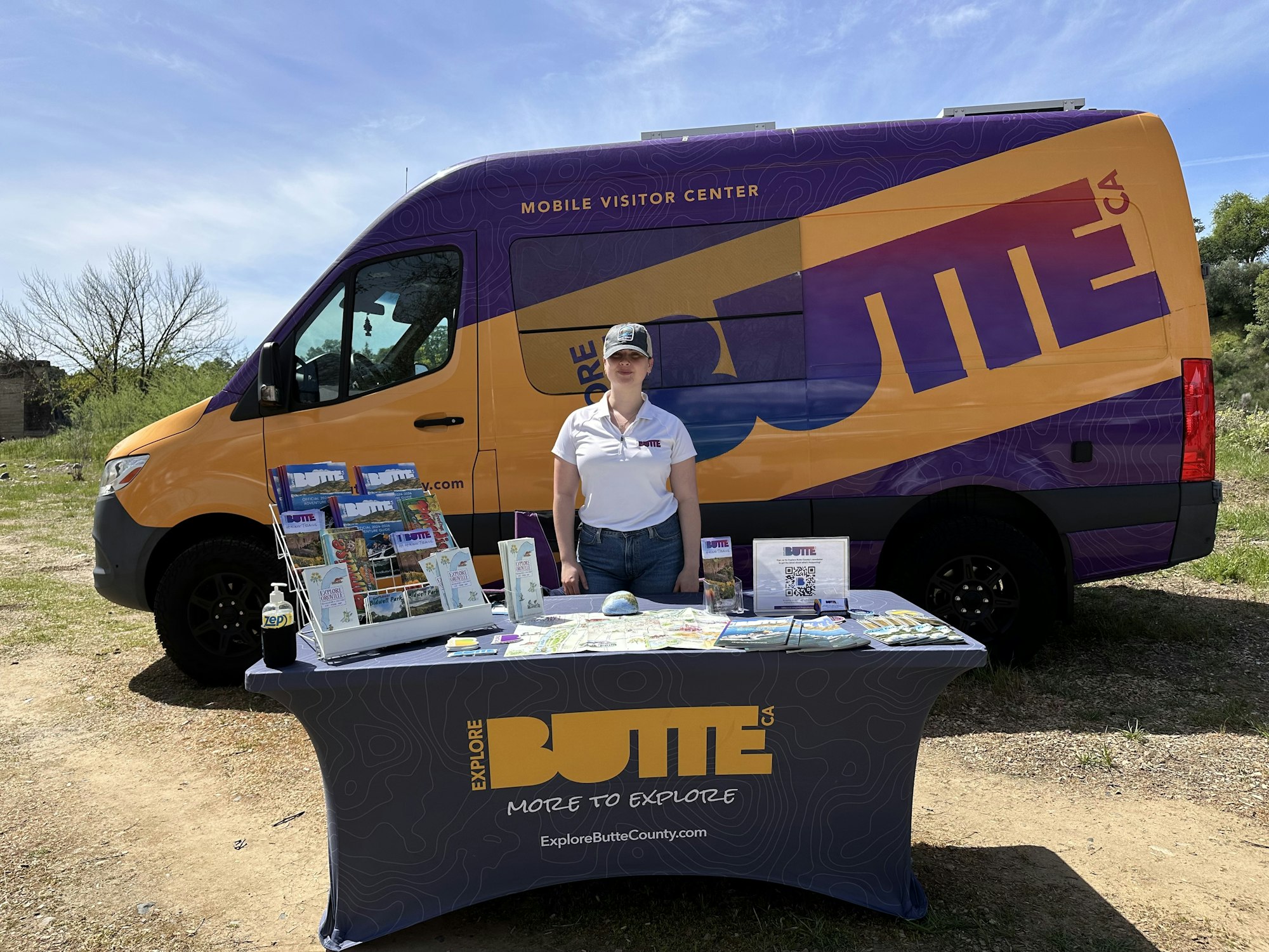 A person stands by a table with brochures and a sign, next to a colorful "Explore Butte County" van.