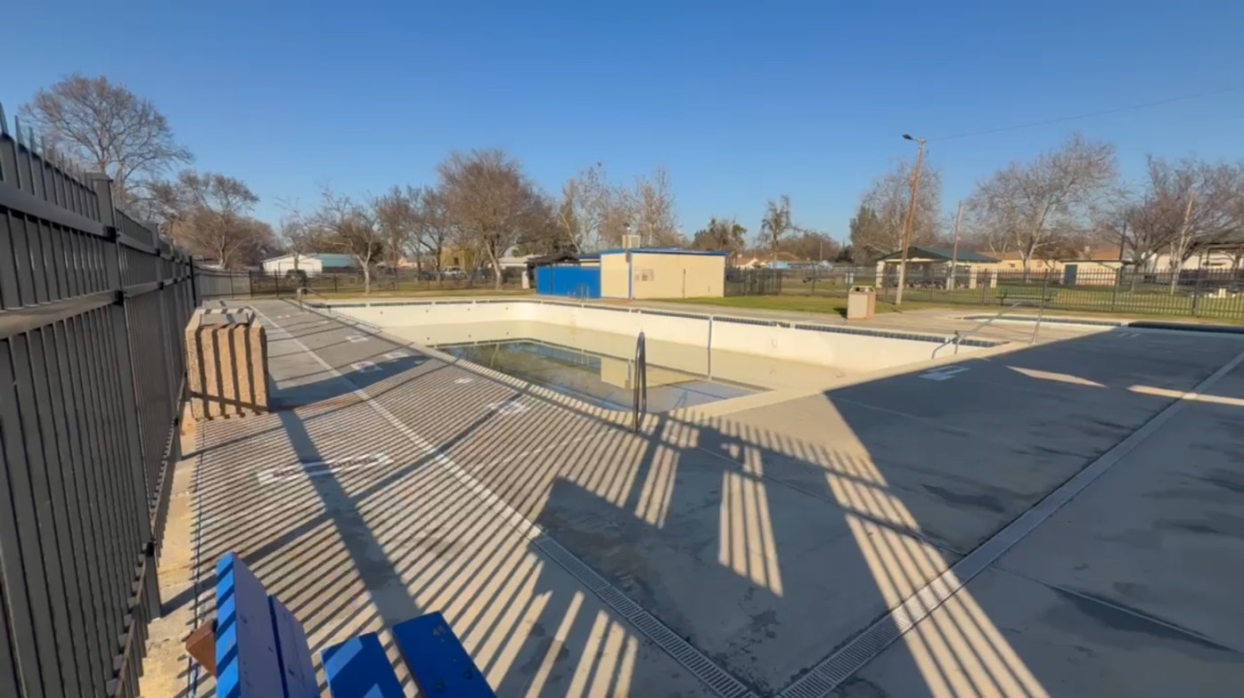 An empty swimming pool surrounded by fences and benches, with a clear blue sky in the background.
